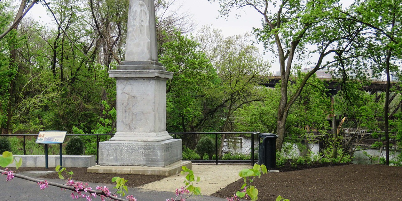 A light gray stone obelisk along the river surrounded by spring foliage